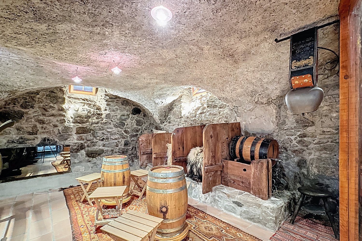 Coin de la cave voûtée avec tonneaux, banc de bois, tapis et cave à vin en arrière-plan.