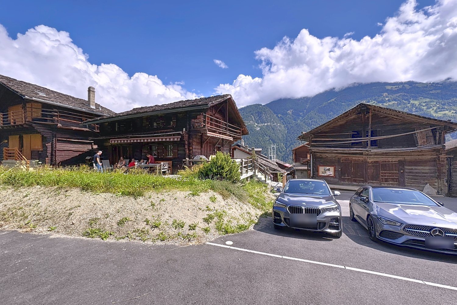 Le chalet et les raccards voisins sous un ciel d'été, façades en madriers sombres, montagnes en arrière-plan.