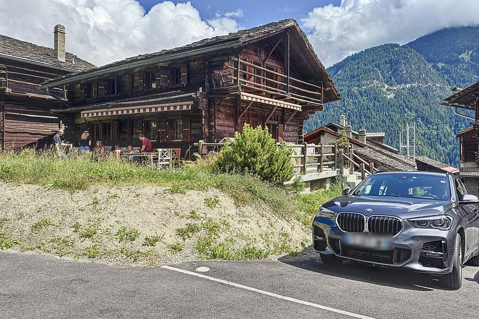 Le chalet vu depuis le chemin : façade en madriers brunis sur socle de pierre, balcon en bois à l'étage, montagnes du Val de Bagnes en arrière-plan.