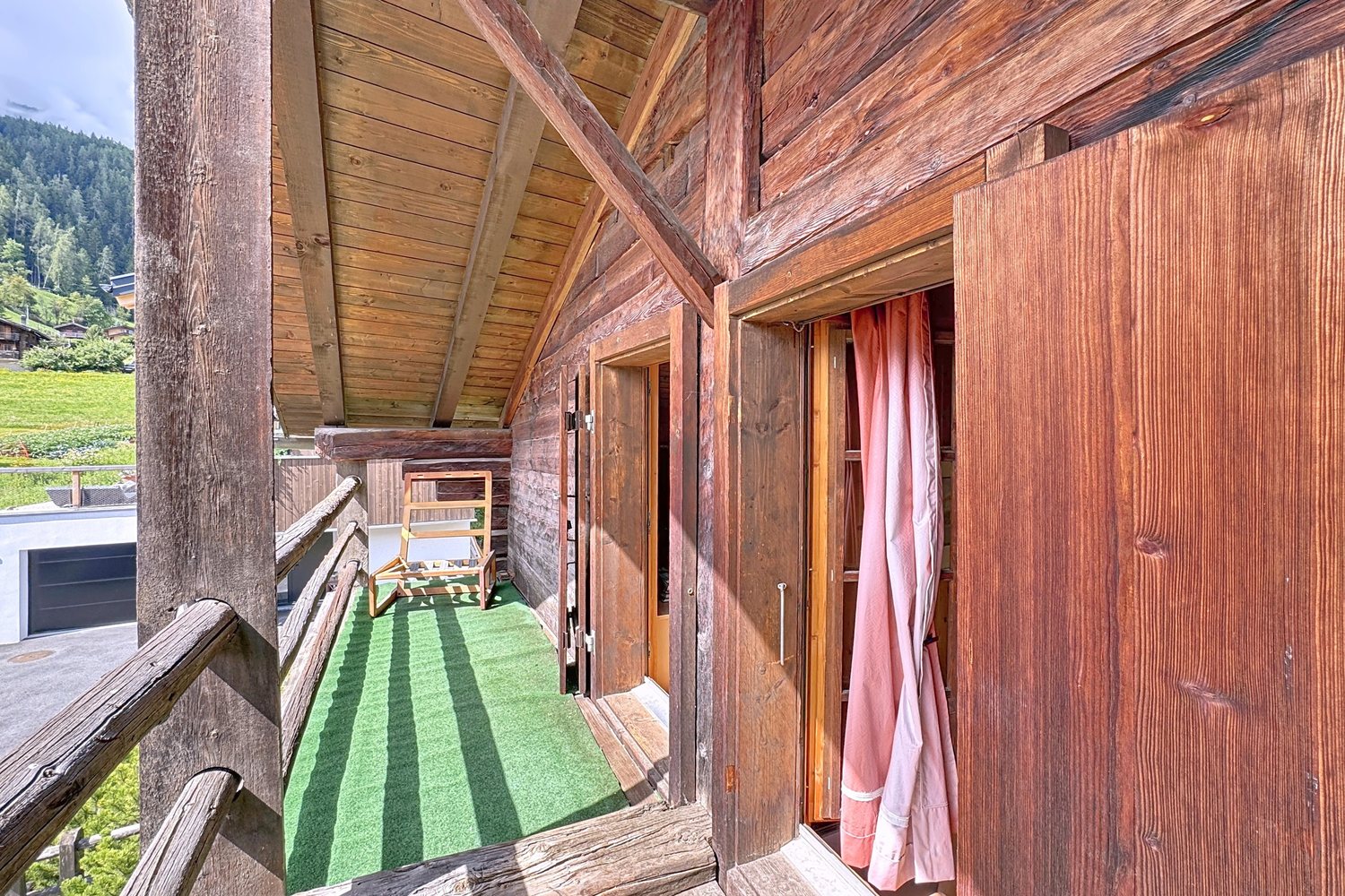 Balcon couvert en bois, tapis vert, chaise longue, avec les prés et les chalets du village en contrebas.